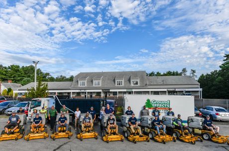 Group of people with yellow lawn mowers in front of a building; blue sky.