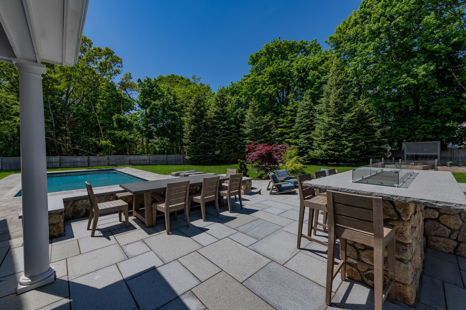 Patio with outdoor kitchen, dining table, chairs, swimming pool, and trees on a sunny day.