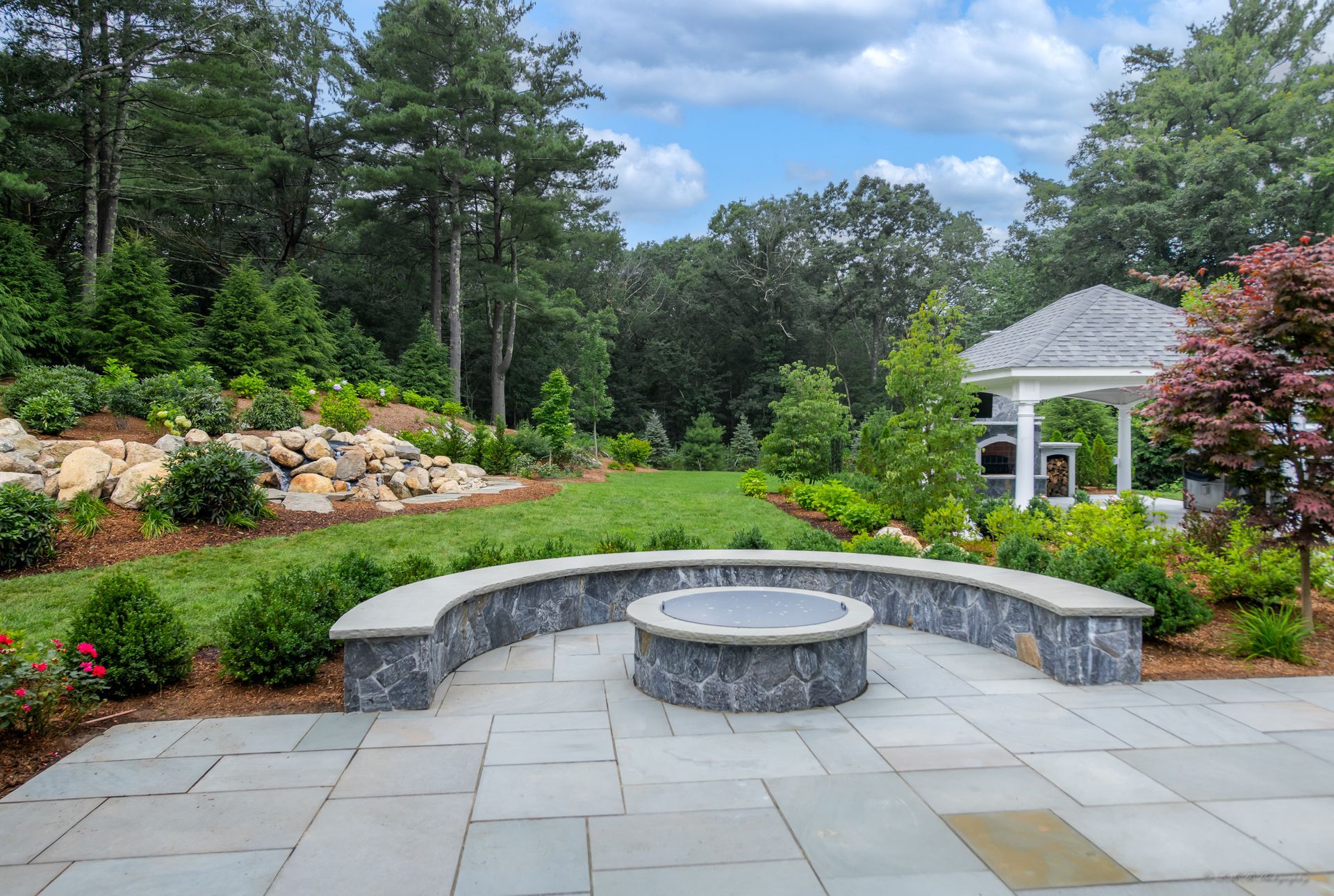 Stone patio with a fire pit, curved seating, and a gazebo in a landscaped backyard.