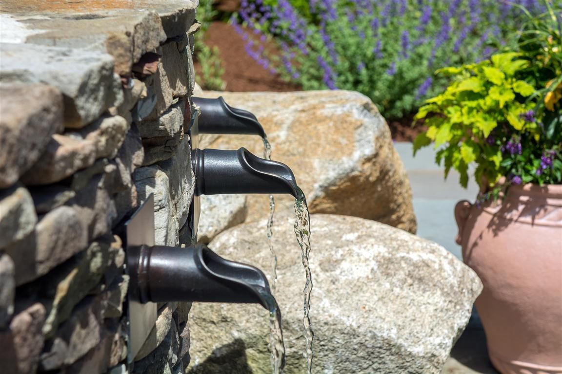 Water flows from three metal spouts in a stone wall into a rock-filled garden with plants.