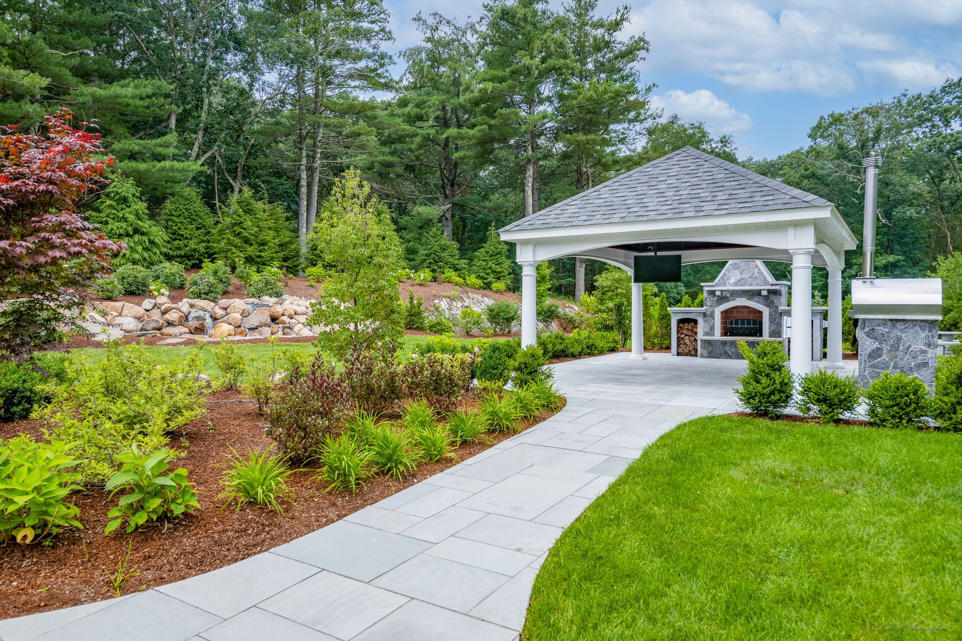 Stone patio with a white gazebo, outdoor fireplace, and manicured landscaping.